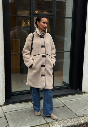 Abrigo de forro polar beige con detalles y botones negros, con un cuello alto y un corte holgado. Combinado con vaqueros acampanados y zapatos beige.