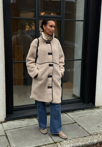 Abrigo de forro polar beige con detalles y botones negros, con un cuello alto y un corte holgado. Combinado con vaqueros acampanados y zapatos beige.