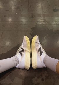 Chaussures de sport blanches avec tige en mesh texturé, semelle intermédiaire jaune, logo noir et semelle plate, associées à des bas gris clair.