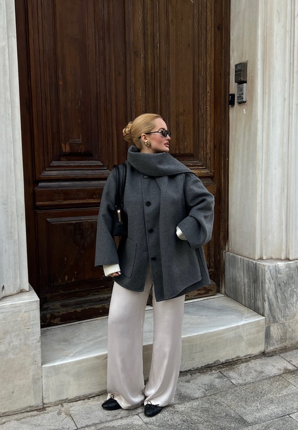 Woman with blonde hair in a bun wears dark grey oversized coat, cream wide-leg trousers, black flats, sunglasses, and gold earrings, standing by a wooden door.