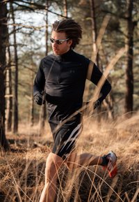 Coureur portant un haut noir à manches longues et un short noir avec des rayures blanches, portant des lunettes de soleil et des chaussures de course avec des accents orange dans un cadre forestier.
