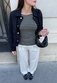 Černá bunda se zlatými knoflíky, pruhovaný černobílý top, krémové plisované kalhoty a malá černá tkaná taška. Černé tenisky s bílými detaily.