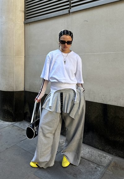 Pantalones de chándal grises holgados con un detalle de falda adjunto, combinados con una camiseta blanca, zapatos amarillos y un bolso negro. Pañuelo negro y gafas de sol.