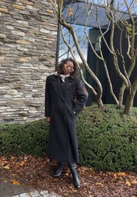 Lungo cappotto nero con design doppiopetto, sciarpa grigia e stivali neri al ginocchio. Sullo sfondo muro di pietra e fogliame verde.