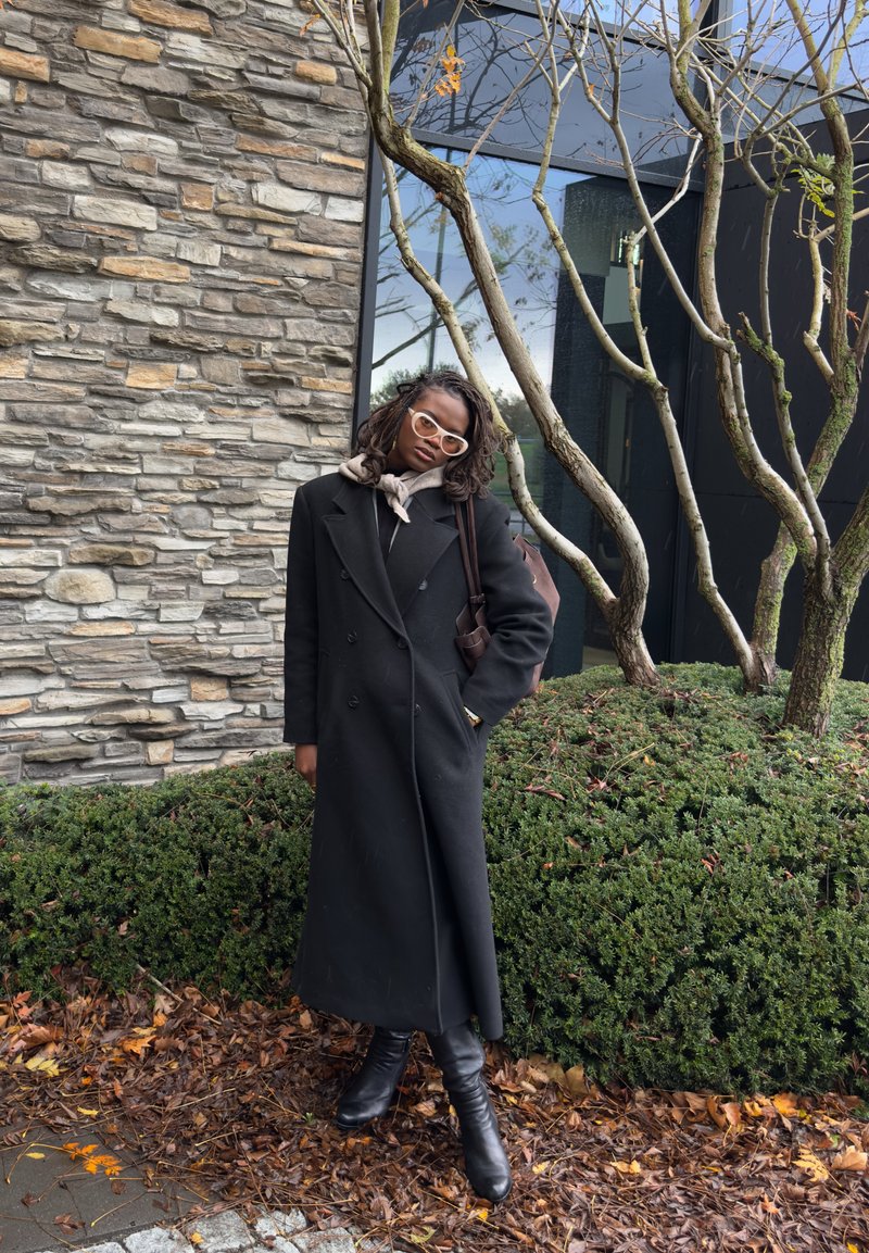 Lång svart rock med dubbelknäppning, en grå halsduk och svarta knähöga stövlar. Bakgrunden har en stenmur och grönska.