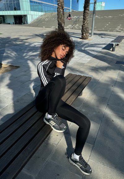 Chaqueta deportiva negra con rayas blancas, leggings negros y zapatillas de running grises. Sentada en un banco de madera con un fondo de azulejos.