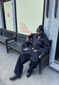 Schwarze Zip-Up-Hoodie, oversized schwarze Hose, schwarze Schuhe mit silbernen Schnürsenkeln. Sitzt auf einer Holzbank neben einer grünen Wand mit Text.