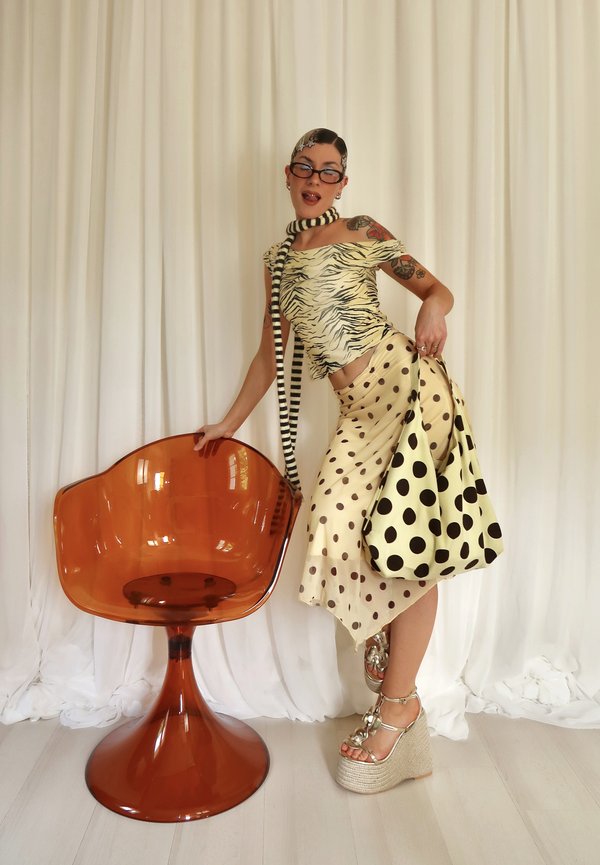 Vrouw met een dierenprint top, een polkadot rok, een gestreepte sjaal en platformsandalen, poserend met een amberkleurige transparante stoel tegen een witte gordijn.