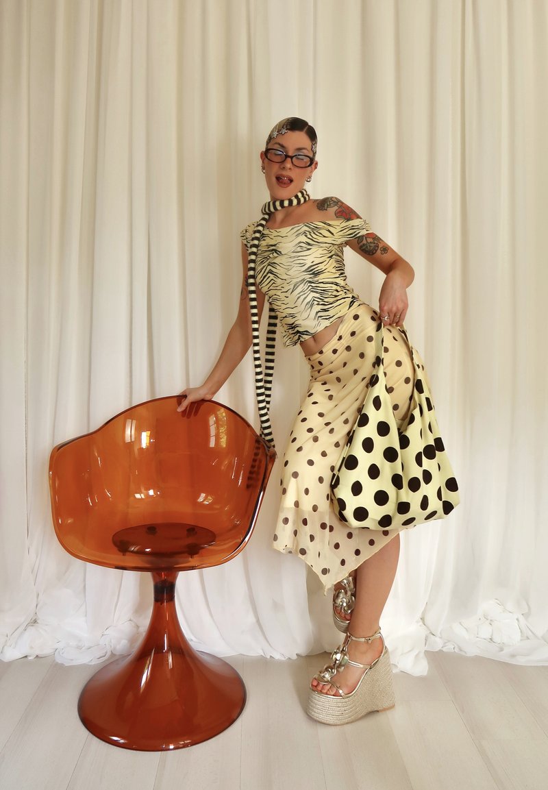 Femme portant un haut imprimé animal, une jupe à pois, une écharpe rayée et des sandales à plateforme, posant avec une chaise transparente ambre devant un rideau blanc.