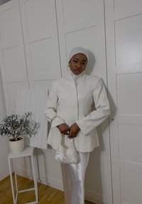 White tailored jacket with a high collar and long sleeves, paired with a silver satin skirt. Holding a small, sequinned white bag.