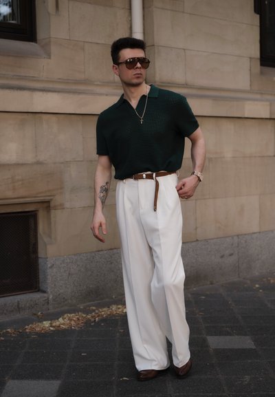 Camisa de polo texturizada verde, pantalones de pierna ancha blancos, cinturón marrón y zapatos marrones. Las gafas de sol y un collar de cadena son accesorios destacados.