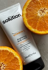 Tubetto bianco di crema corpo illuminante con vitamina C, con testo nero, circondato da due arance tagliate a metà. Gocce d'umidità sulla superficie della crema.