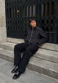 Giacca nera zip-up, pantaloni neri larghi e stivali neri lucidi. Seduto su gradini di pietra con sfondo in granito grigio e griglie di metallo.