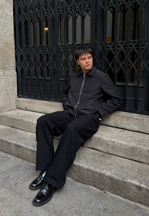 Giacca nera con zip, pantaloni neri larghi e stivali neri lucidi. Seduto su gradini di pietra con uno sfondo in granito grigio e griglie metalliche.