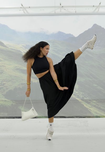 Top corto negro y falda larga en movimiento, complementada con pulseras doradas y un bolso blanco. Calzado: zapatos blancos con un diseño único.