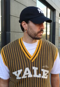 Brown and yellow striped knitted vest with "YALE" in white letters, worn over a white T-shirt; paired with a black cap featuring a logo.