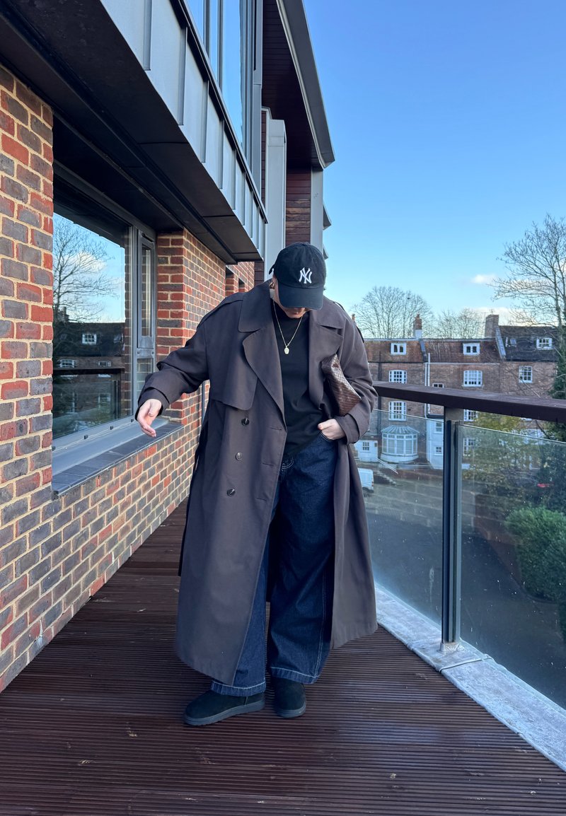 Personne portant un long manteau sombre, un jean large, une casquette noire et des bottes, debout sur un balcon à côté d'un bâtiment en brique sous un ciel bleu clair.