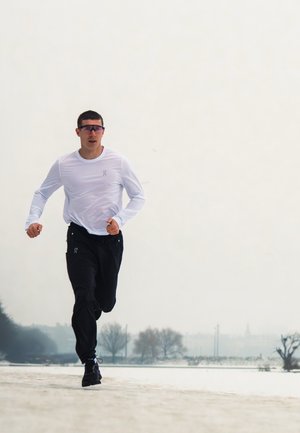 Hombre con gafas de sol, camisa blanca de manga larga y pantalones negros corriendo al aire libre en un día con niebla cerca de árboles desnudos.