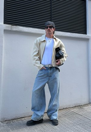 Hombre con chaqueta beige, camisa blanca, vaqueros azules holgados, zapatos negros, gorra gris, gafas de sol, sosteniendo bolso negro, de pie contra una pared blanca al aire libre.