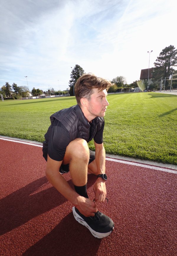 Hombre con ropa deportiva negra agachado en la pista de atletismo al aire libre atándose el zapato, con campo de césped verde y árboles de fondo.