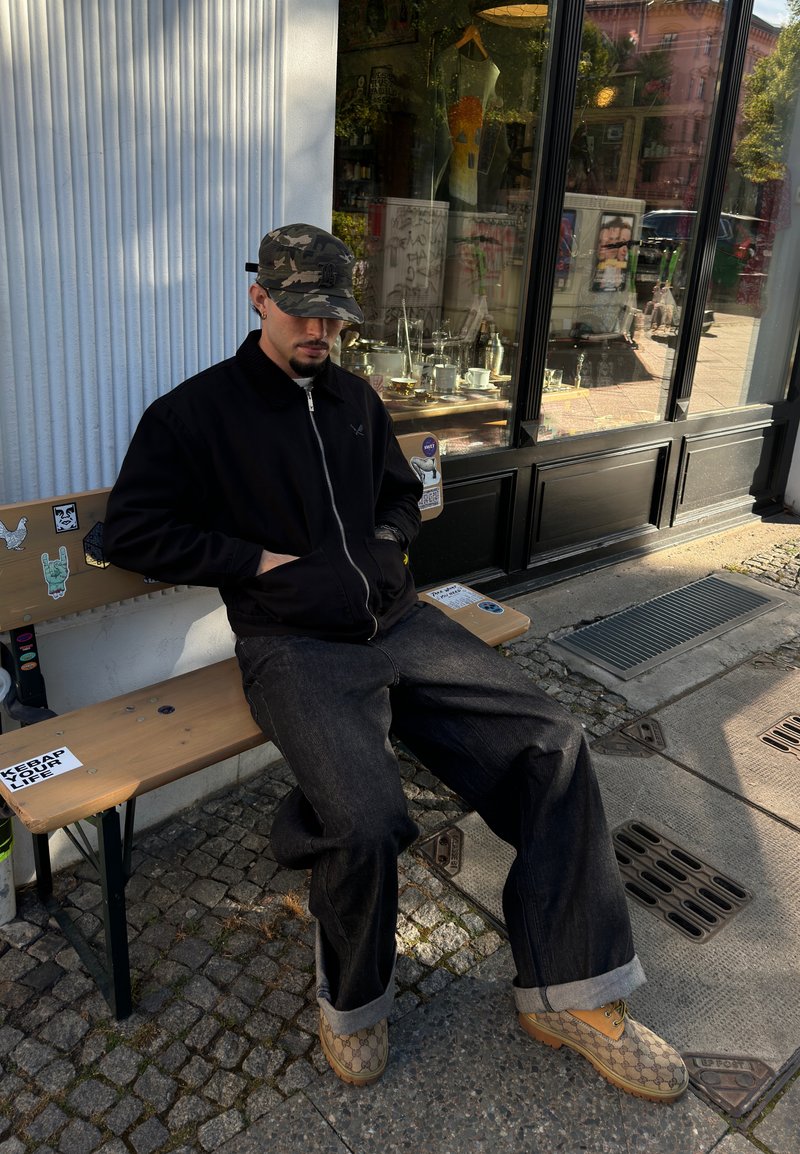 Μαύρο φερμουάρ μπουφάν, σκούρες φαρδιές τζιν με διπλωμένα μπατζάκια και καφέ παπούτσια με σχέδιο Gucci. Καθισμένος σε μια ξύλινη παγκάκι έξω.