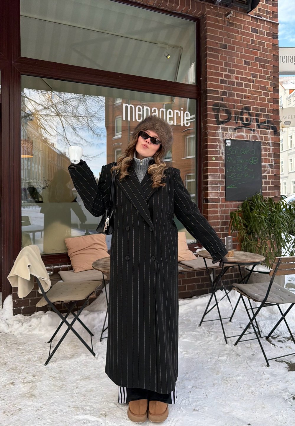 Personne en manteau noir à fine rayures et chapeau en fourrure tenant une tasse blanche à l'extérieur d'un café enneigé avec des tables et des chaises en bois.