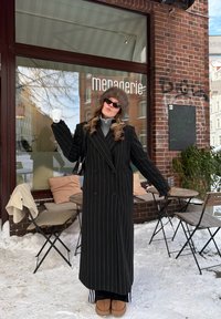 Personne en manteau noir à fine rayures et chapeau en fourrure tenant une tasse blanche à l'extérieur d'un café enneigé avec des tables et des chaises en bois.
