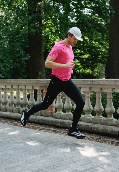 La ropa de running incluye una camiseta de mangas cortas de color rosa brillante y leggings ajustados negros. La figura lleva zapatillas de running en blanco y negro y una gorra blanca.