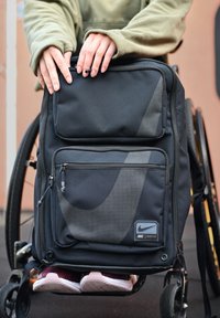 Schwarzer Rucksack mit strukturiertem Oberflächen und vorderer Netztasche. Verfügt über zwei Reißverschlüsse, Nike-Logo und steht auf einem Rollstuhl, während Sneaker sichtbar sind.