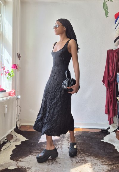 Mujer con vestido negro sin mangas y zuecos negros con plataforma, sosteniendo un pequeño bolso negro, de pie en una habitación iluminada por el sol cerca de una ventana.