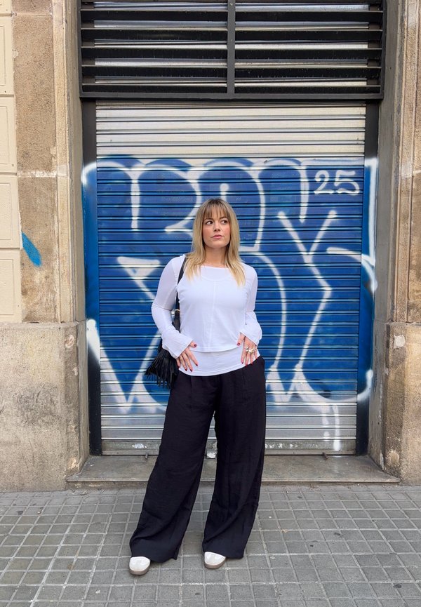 Donna con capelli biondi, indossa una maglietta bianca e pantaloni neri larghi, in piedi con le mani in tasca davanti a una saracinesca di metallo coperta di graffiti blu.