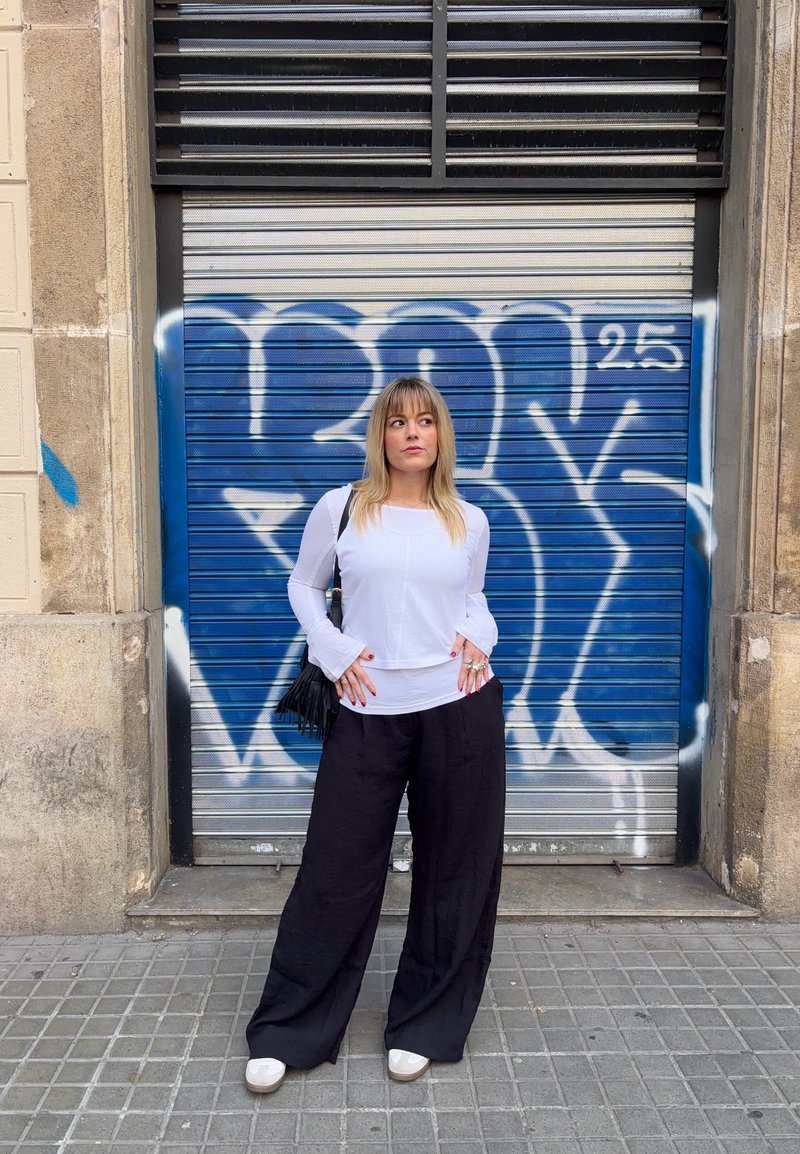 Szőke hajú nő fehér felsőben és fekete, bő szárú nadrágban áll zsebeiben tartott kézzel egy kék graffiti által borított fém redőny előtt.