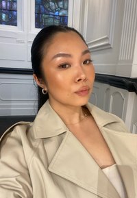 Abrigo trench beige con cuello muesca y hombreras. Textura suave, llevado sobre una blusa clara. Pelo corto y aretes de aro visibles.
