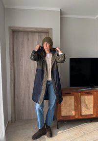 Femme portant un manteau vert et bleu marine, un pull beige, un jean bleu, des bottes marron et un bonnet en tricot olive, à l'intérieur près d'une porte en bois et d'un meuble TV.