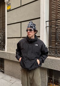 Άτομο με μαύρο μπουφάν North Face, μαύρο και λευκό σκουφάκι με σχέδιο, γυαλιά ηλίου, στέκεται σε πεζοδρόμιο της πόλης κοντά σε πέτρινο τοίχο και σιδερένια περίφραξη.