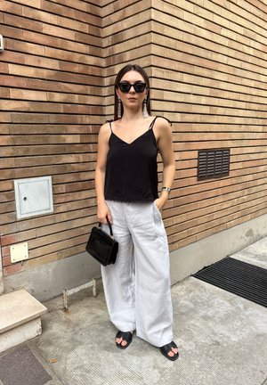 Camiseta sin mangas negra con encaje, pantalones anchos blancos, sandalias negras y un pequeño bolso negro. El fondo presenta una pared de ladrillo.