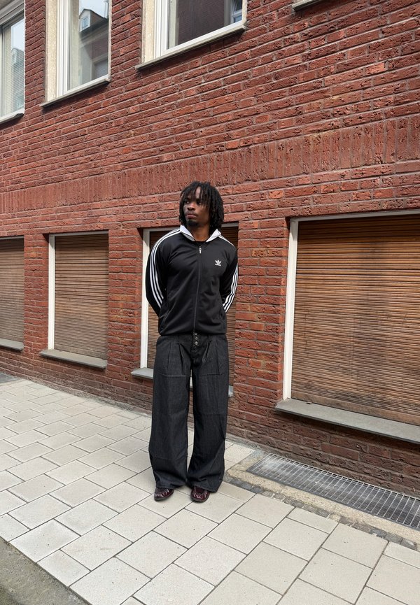 Hombre con rastas, vestido con una chaqueta negra de Adidas y pantalones anchos, está de pie en la acera junto a un edificio de ladrillo rojo con persianas de madera cerradas.