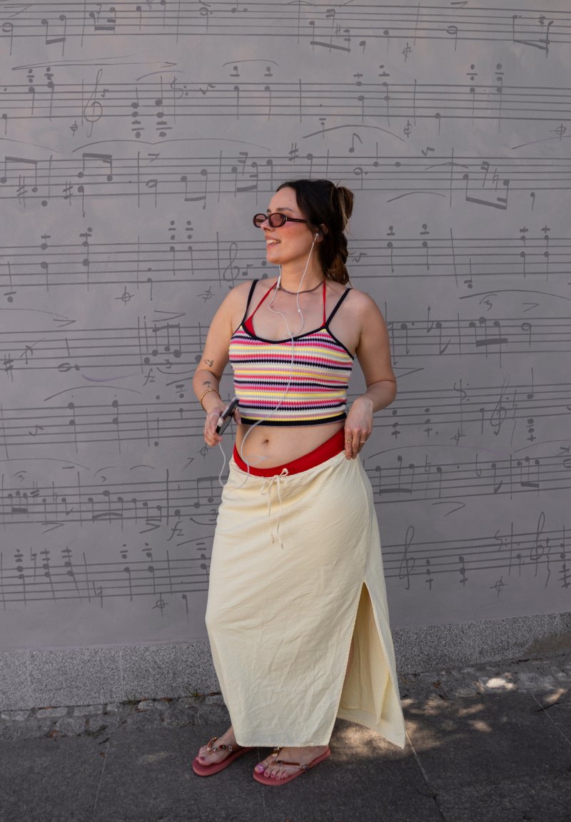 Jonge vrouw met zonnebril, gekleed in een gestreept crop top en een lange rok, staat glimlachend met oortjes en smartphone bij een muurschildering met een muzieknoot.