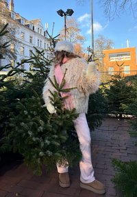 Cream faux fur jacket, pink scarf, white gloves, light-coloured trousers, and brown boots; surrounded by green Christmas trees in an outdoor setting.