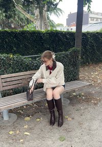 Kremfarget jakke med brun krage, rød topp, korte denimshorts, brune knelengde støvler, sitter på en trebenk i en park.