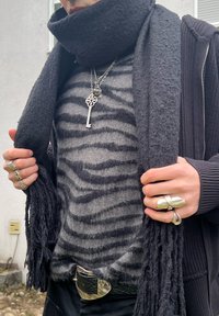 Personne portant une écharpe noire, un pull gris à rayures, un collier avec une clé en argent, plusieurs bagues en argent et une ceinture noire avec une grande boucle en argent.
