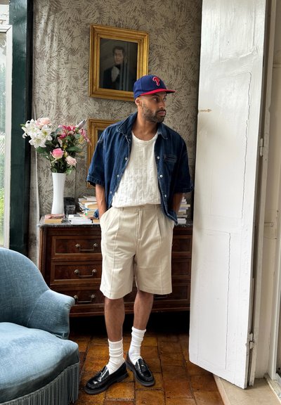 Chaqueta vaquera, camisa blanca de lino y pantalones cortos beige. Mocasines negros con calcetines blancos. Escenario interior con arreglo floral y muebles de madera.