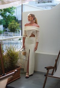 Kreem värvuses off-white kudumiga lühike topp, millel on õlavarre alt avatud disain, koos kõrge lõhikuga, sobiva kreemivärvi maxiseelikuga; hoiab käes väikest ümmargust kotti.