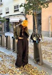 Čierne široké nohavice s vyšívaným vzorom, zelená bomber bunda, oranžová čiapka s logami, vrstvené náhrdelníky a čierna kabelka.