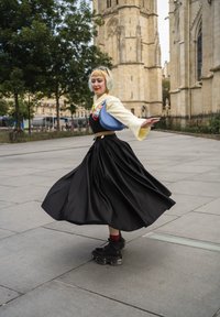Persona con el cabello teñido girando en una falda negra, blusa amarilla, bolso azul al hombro y botas gruesas negras en una calle de la ciudad cerca de edificios históricos.