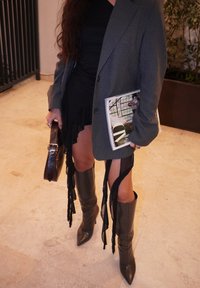 Szary blazer w prążki w oversize'owym kroju, czarna sukienka z frędzlami, czarne kozak do kolan na szpilce, trzymająca czarną torebkę i magazyn.