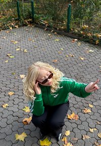 Casaco verde com fecho éclair e riscas brancas, calças pretas e óculos de sol. Cabelo loiro ondulado. Piso pavimentado com folhas amarelas dispersas.