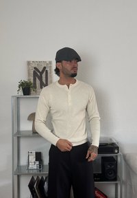 Cream-coloured ribbed long-sleeve henley shirt with buttons, teamed with black trousers. A grey flat cap and tattoos are visible on the arms, with a modern shelf in the background.