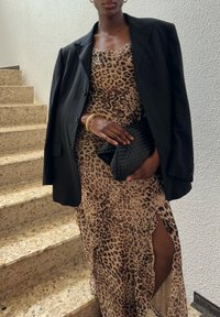 Vestido de estampa de leopardo com detalhes em babados, acompanhado por um blazer oversized preto, segurando uma clutch preta de vime, em frente a um fundo de escadaria.