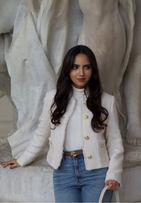 Giacca bianca con texture e bottoni dorati, indossata sopra un dolcevita beige e jeans azzurri. Tiene una borsa bianca contro uno sfondo in marmo.